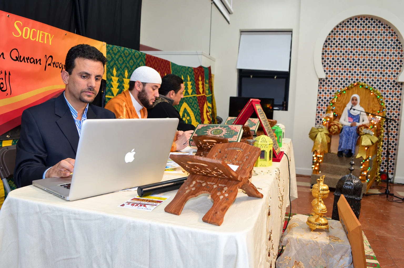 Al Huda community members at a Quran event, with books and wooden Quran stands on the table.